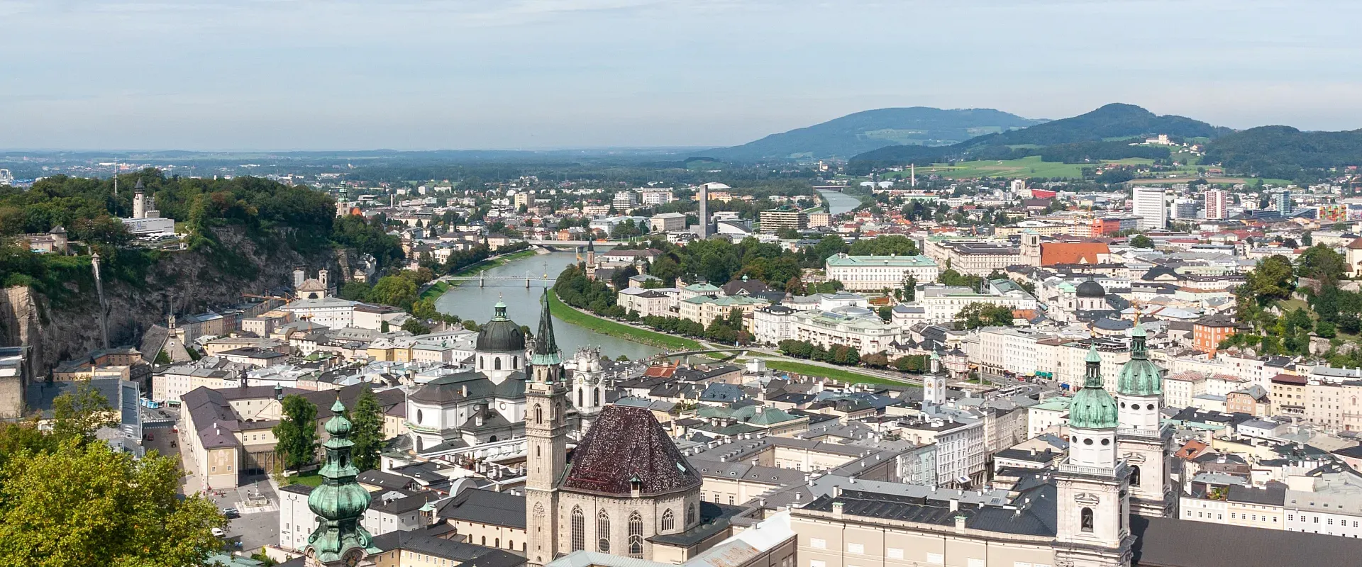 Panoramablick auf die Stadt Salzburg, mit der Salzach und den Alpen im Hintergrund, grünen Hügeln und historischen Gebäuden der Altstadt im Vordergrund.