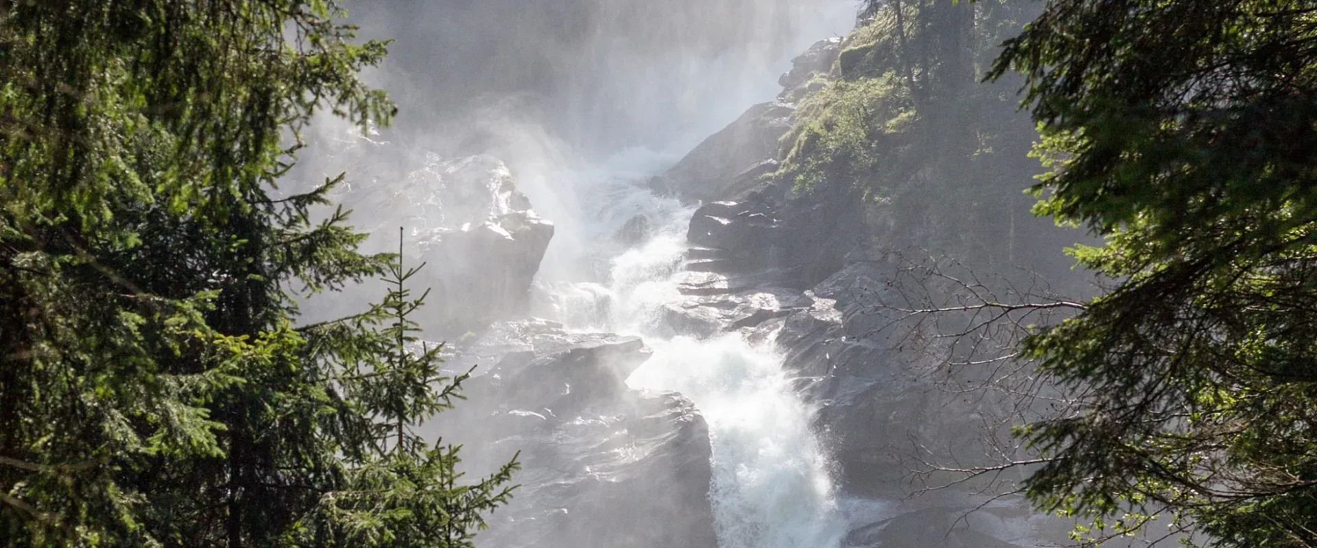Ein beeindruckender Wasserfall stürzt zwischen bewaldeten Felsen herab, umgeben von dichtem Grün und angestrahlt von Sonnenlicht, das durch die Bäume fließt.