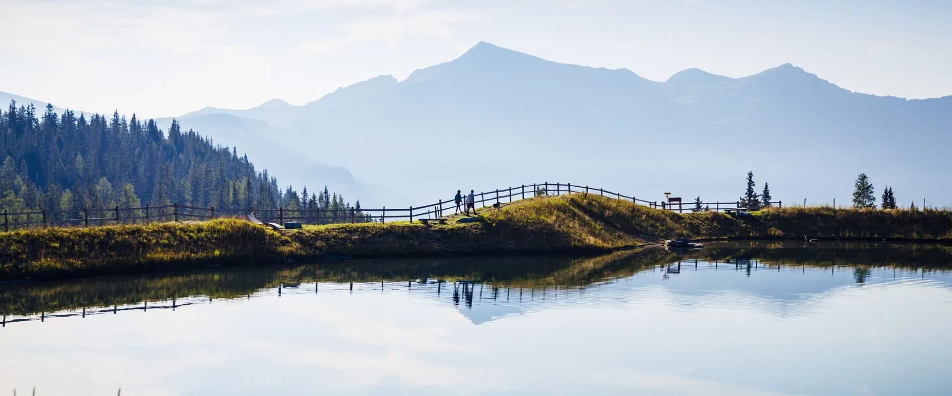 Ein idyllischer See mit klarem Wasser, das die umliegenden Berge und einen blauen Himmel spiegelt. Ein hölzerner Zaun verläuft entlang des Ufers.
