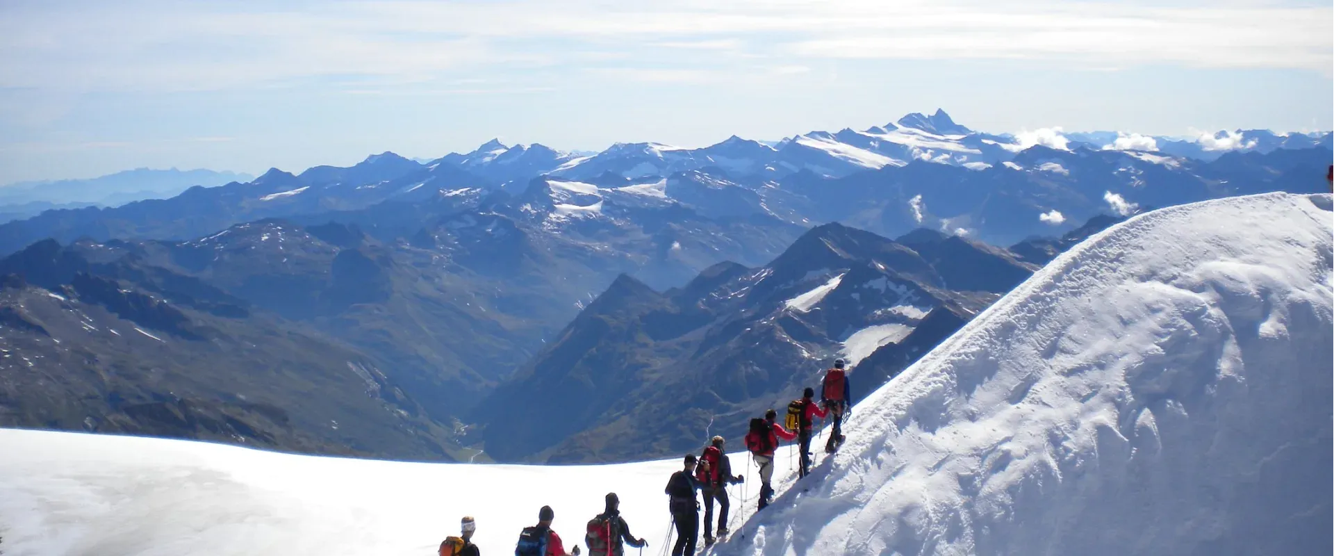Eine Gruppe von Menschen wandert in einer Schneelandschaft mit Berggipfeln im Hintergrund unter klarem Himmel. Die Sonne scheint auf den Schnee.