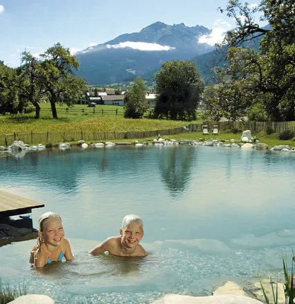 Zwei lachende Kinder mit blonden Haaren schwimmen in einem natürlichen Teich. Im Hintergrund ist eine grüne Wiese, Bäume und ein Bergpanorama zu sehen.