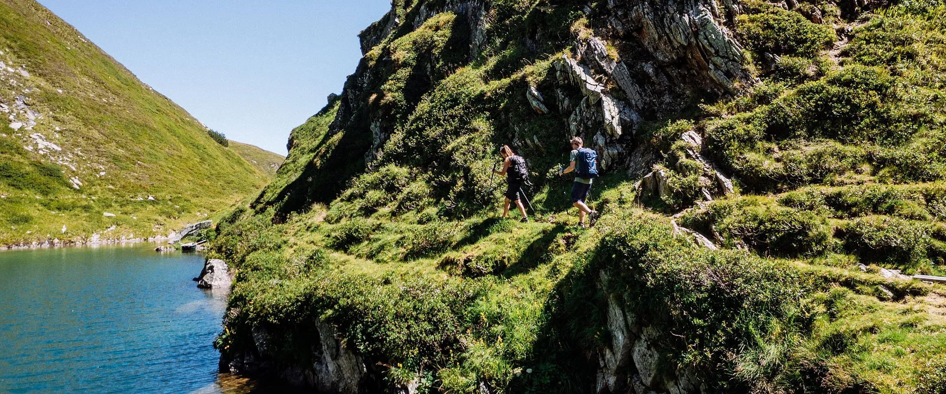 Zwei Wanderer gehen entlang eines schmalen, grasbewachsenen Pfades neben einem klaren, blauen Bergsee, umgeben von steilen, grünen Hügeln bei sonnigem Wetter.