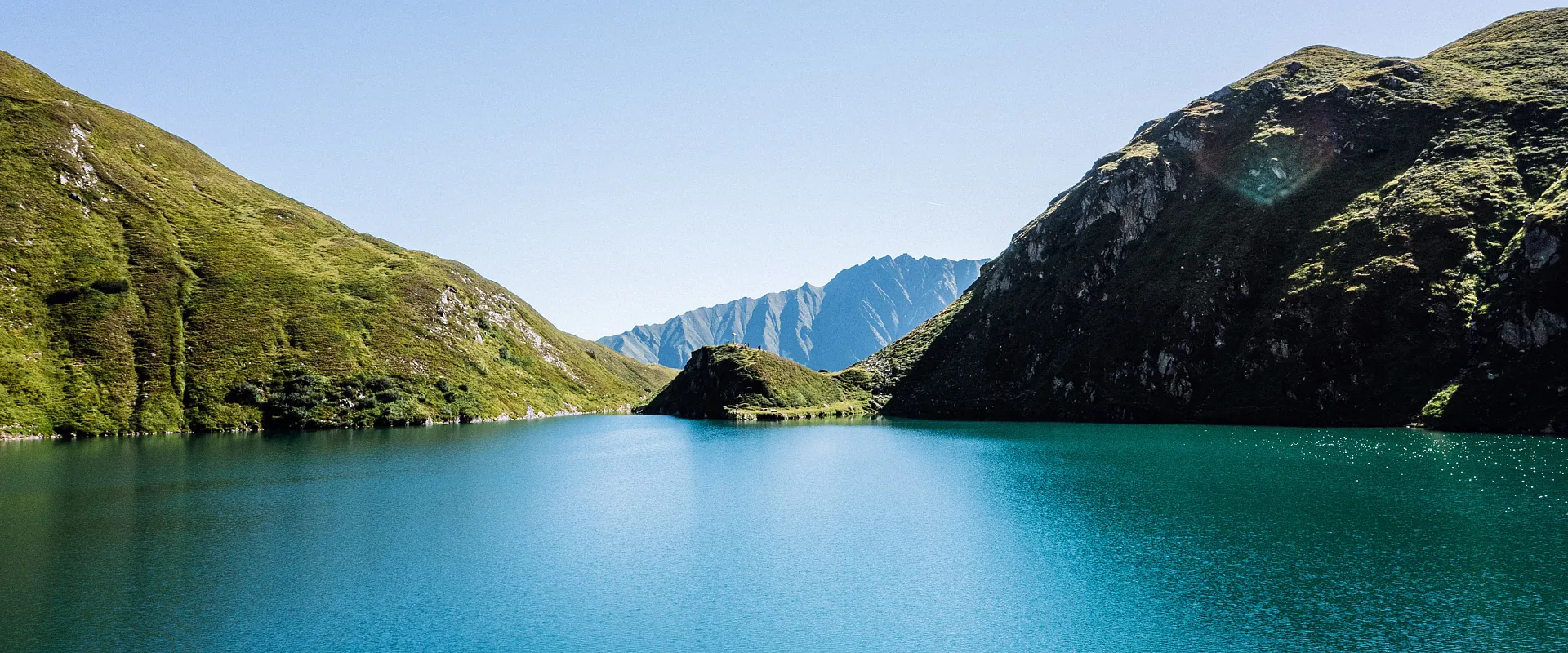 Ein türkisblauer Bergsee umgeben von grünen, bewachsenen Hügeln und höheren, steilen Gipfeln im Hintergrund unter klarem, blauem Himmel.