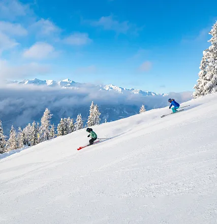 Zwei Menschen fahren in Wintersportkleidung auf einer schneebedeckten Piste. Umgeben von verschneiten Bäumen und Bergen unter blauem Himmel. Es ist sonnig.