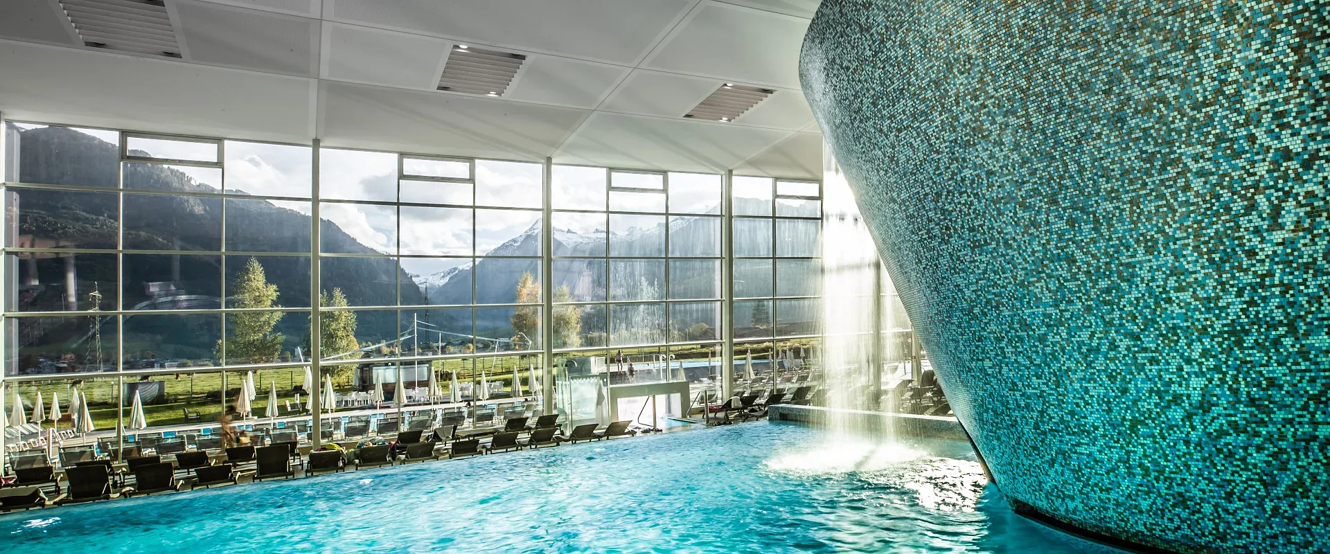 Schwimmbad mit glitzerndem blauen Wasser in einem modernen Gebäude. Große Fenster bieten Blick auf bewaldete Berge unter einem bewölkten Himmel.