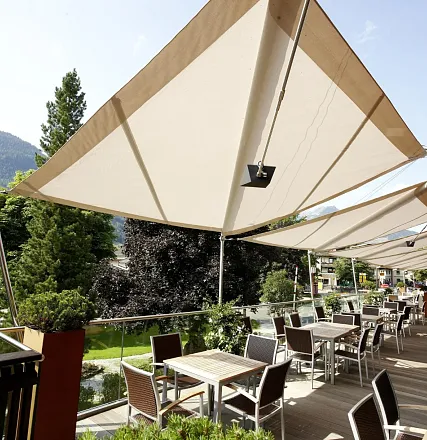 Sonnensegel spenden Schatten auf einer Terrasse mit Tischen und Stühlen, umgeben von grünen Bäumen und einem Blick auf eine malerische Berglandschaft.