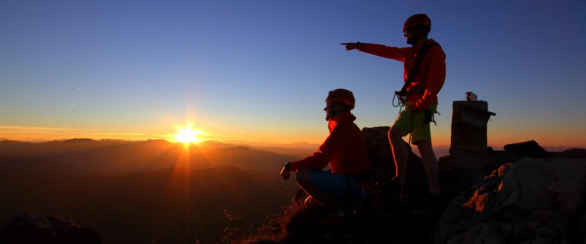Zwei Kletterer beobachten den Sonnenaufgang von einem Berggipfel. Einer zeigt in die Ferne. Der Himmel ist klar, und die Sonne taucht alles in orangefarbenes Licht.