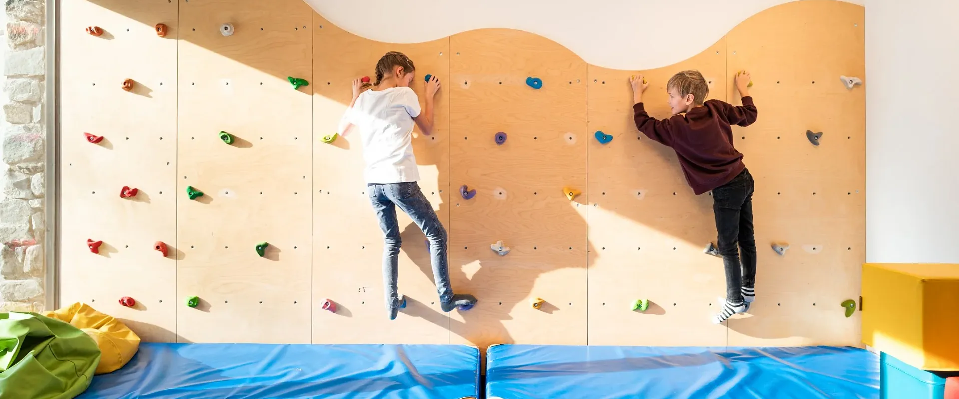 Zwei Kinder klettern an einer bunten Indoor-Kletterwand, während eine weiche Matte den Boden schützt. Die Szene wirkt lebendig und verspielt.