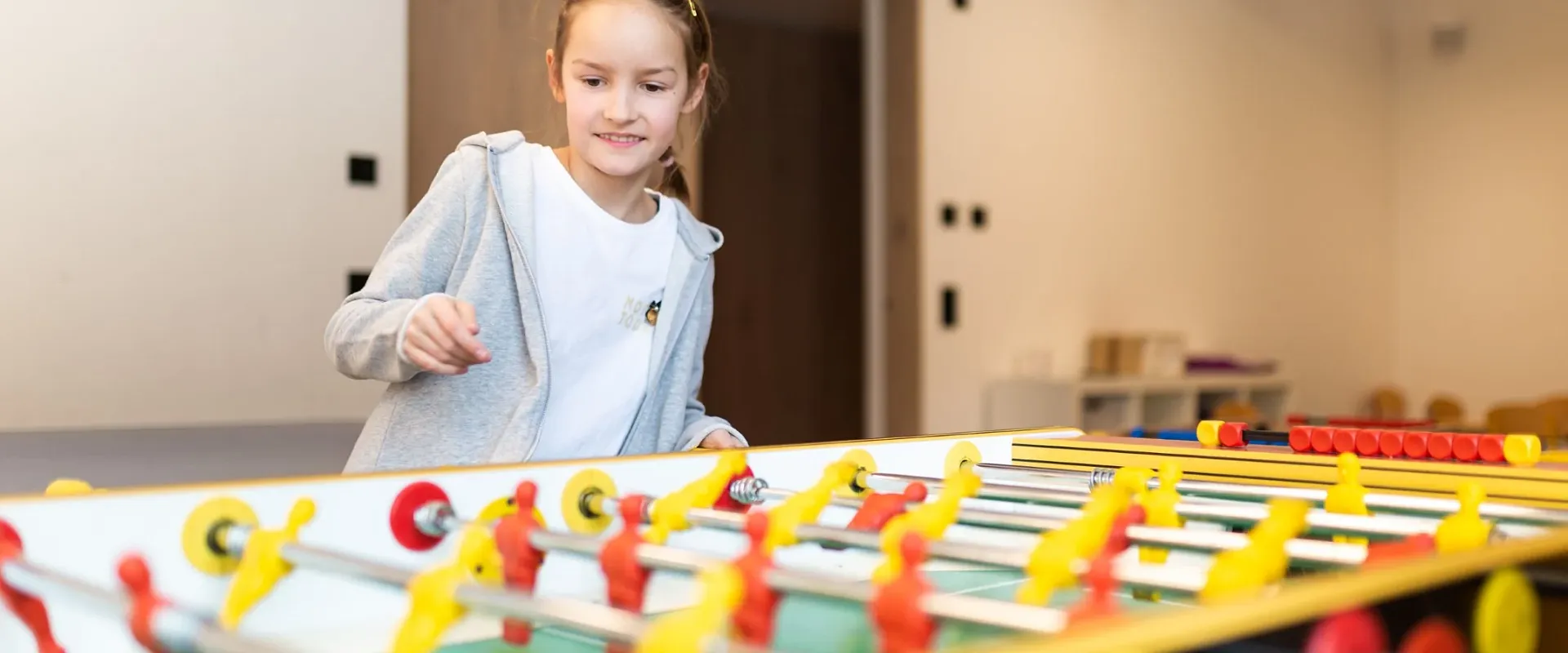 Ein Kind spielt Tischfußball in einem Raum. Das Kind trägt einen grauen Pullover und konzentriert sich auf das Spielfeld, das bunt gestaltet ist.