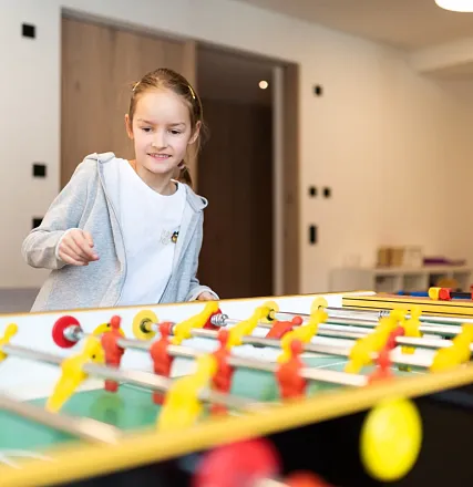 Mädchen spielt fröhlich an einem Tischkicker. Die Spielfiguren sind in Rot und Gelb, und der Raum wirkt modern und gut beleuchtet.