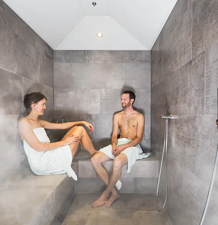 Zwei Personen sitzen entspannt in einem dampfenden Dampfbad. Beide tragen Handtücher und führen ein Gespräch. Der Raum ist in neutralen Grautönen gehalten.