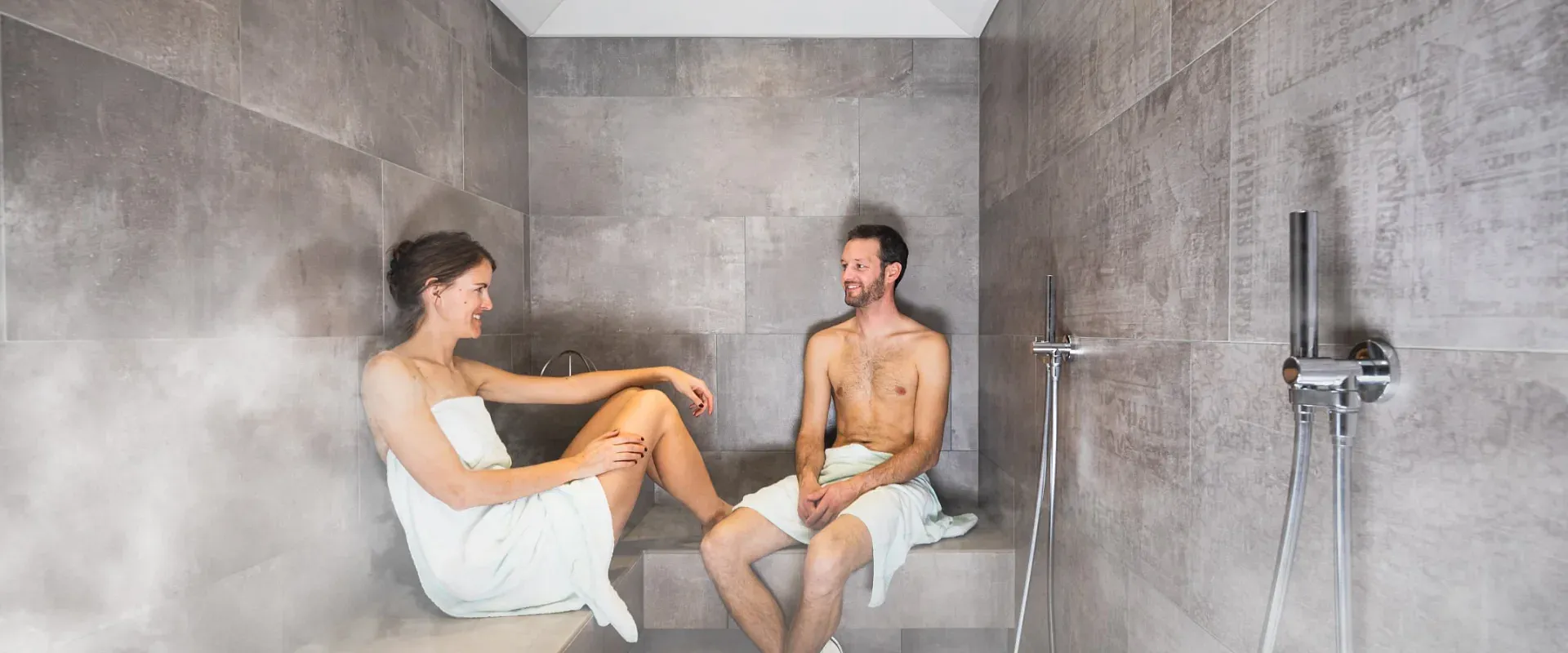 Zwei Personen entspannen in einem modernen Dampfbad mit grauen Fliesenwänden. Sie sind in Handtücher gehüllt und sitzen auf Bänken, umgeben von Dampf.