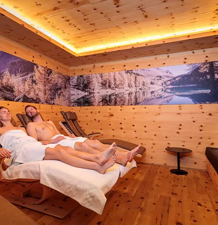 Zwei Personen entspannen in einem Wellnessraum mit Holzwänden. Sie liegen auf Liegestühlen, umringt von beruhigenden Bildern von Natur und Bergen.