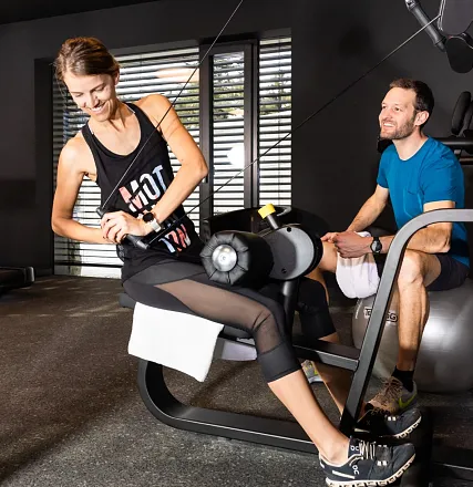 Zwei Personen trainieren im Fitnessstudio. Eine Frau benutzt eine Maschine für Beinübungen, während ein Mann in Sportkleidung daneben sitzt und lächelt.