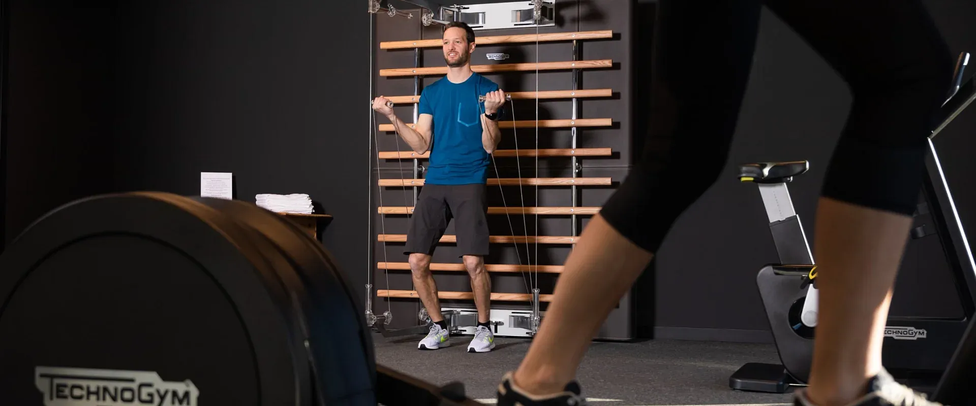 Ein Fitnessstudio mit moderner Ausstattung. Im Fokus sind die Beine einer Person auf einem Crosstrainer zu sehen, während jemand im Hintergrund Übungen macht.