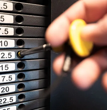 Nahaufnahme einer Hand, die ein Gewicht an einem Fitnessgerät verstellt. Die Gewichtsplatte ist mit verschiedenen Zahlen beschriftet, die das Gewicht angeben.