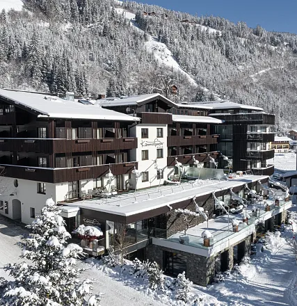 Großes, modernes Alpenhotel im Winter, umgeben von verschneiten Bergen. Dunkle Holzfassaden und Terrassen mit Glasgeländern unterstreichen die Architektur.