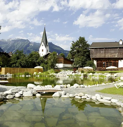 Idyllische Landschaft mit einem Teich im Vordergrund, umgeben von Steinen. Eine Kirche mit hohem Turm und traditionelle Häuser vor einer Bergkulisse.