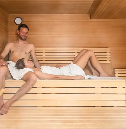 Ein Paar entspannt in einer Sauna. Der Mann sitzt, während die Frau mit einem Handtuch bekleidet ihren Kopf auf seinen Schoß legt. Holzwände umgeben sie.