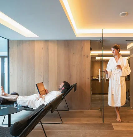 Ein Mann liegt entspannt auf einer Liege mit einem Laptop, während eine Frau im Bademantel durch eine Glastür das Zimmer betritt. Modernes, helles Design.