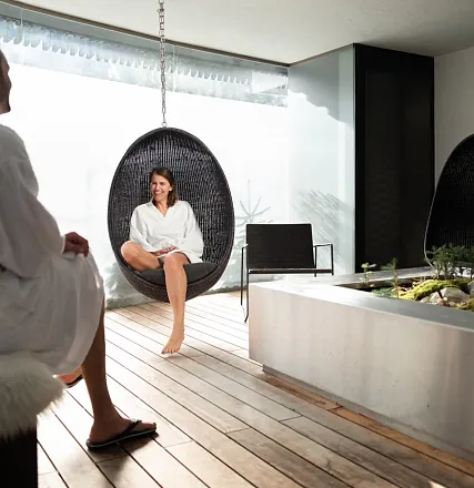 Personen in einem Wellnessbereich; eine Frau sitzt entspannt in einem hängenden Stuhl, in Bademänteln gekleidet. Der Raum ist modern und hell gestaltet.