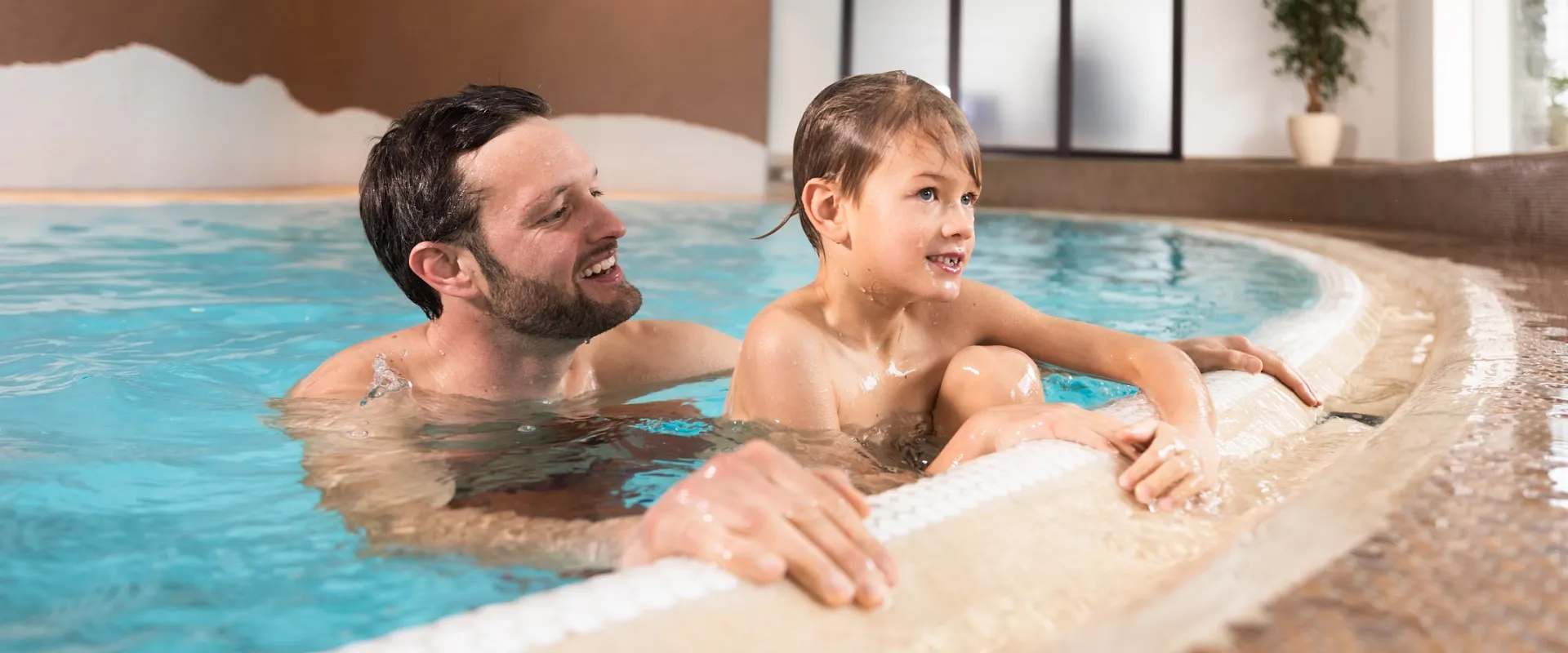 Ein Mann und ein Kind genießen zusammen Schwimmzeit in einem Hallenbad. Sie lachen und scheinen sich auf dem Beckenrand zu entspannen.
