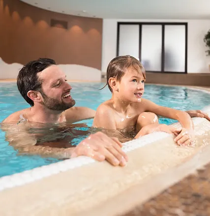 Ein lachender Mann und ein Kind schwimmen in einem Innenpool. Das Kind sitzt am Rand, während der Mann daneben im Wasser steht. Beide wirken entspannt und fröhlich.