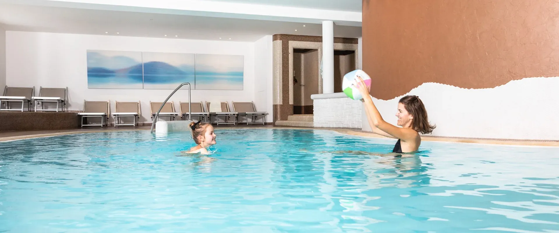 Eine Frau spielt mit einem Kind im Innenpool, wirft einen bunten Ball. Im Hintergrund sind Liegestühle und ein Wandbild zu sehen.