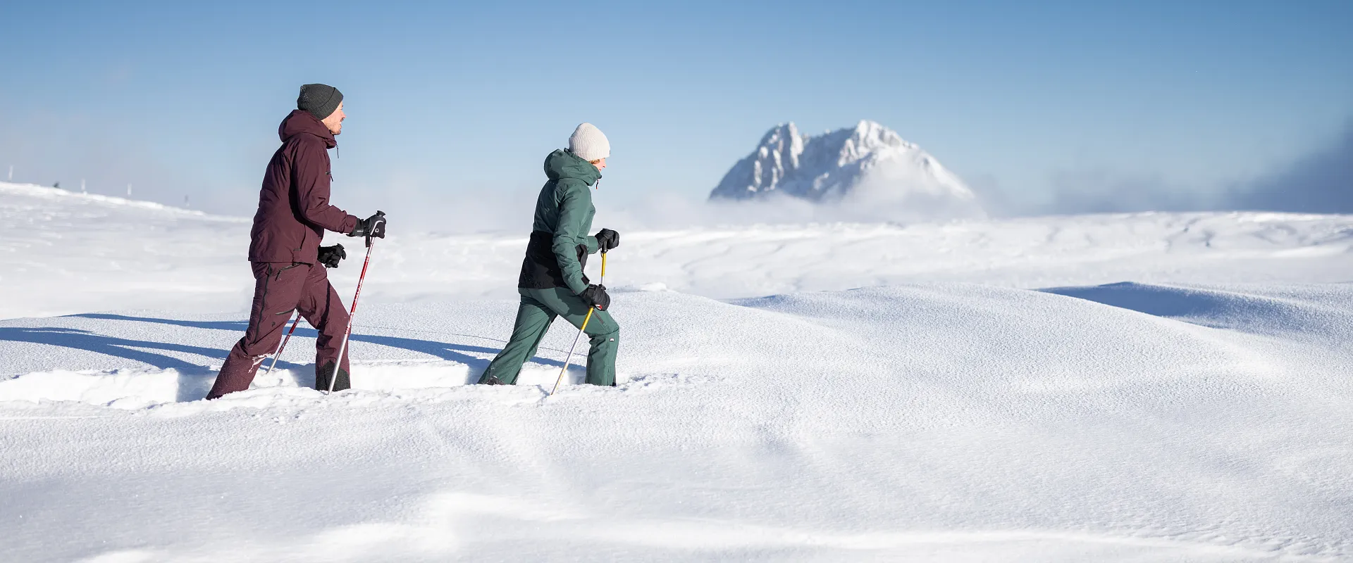 Zwei Menschen wandern in Winterkleidung über eine schneebedeckte Landschaft, mit einem Berg im Hintergrund unter klarem blauem Himmel.