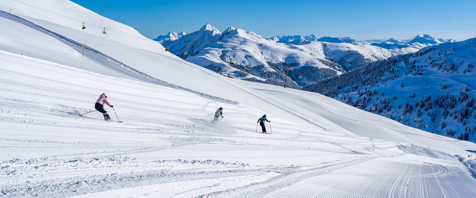 Menschen fahren auf frisch präparierten Pisten Ski, umgeben von schneebedeckten Bergen und blauem Himmel. Die Winterlandschaft wirkt friedlich und idyllisch.
