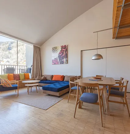 Helles Wohnzimmer mit großen Fenstern, einer Couch mit bunten Kissen, einem Holzesstisch und modernen Stühlen. Ein Gemälde hängt an der Wand.