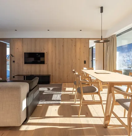 Modernes Wohnzimmer mit Holzwänden, einem großen Tisch und einem Sofa. Ein Fernseher hängt an der Wand. Durch ein Fenster fällt helles Tageslicht ein.