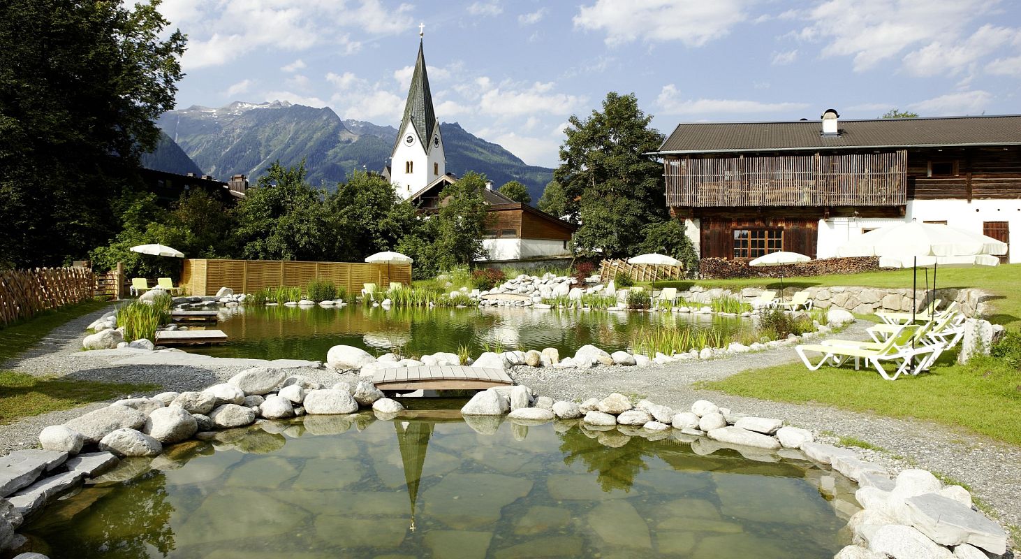 Eine idyllische Landschaft mit einem Teich im Vordergrund, umgeben von Liegestühlen. Im Hintergrund steht eine Kirche vor malerischen Berggipfeln.