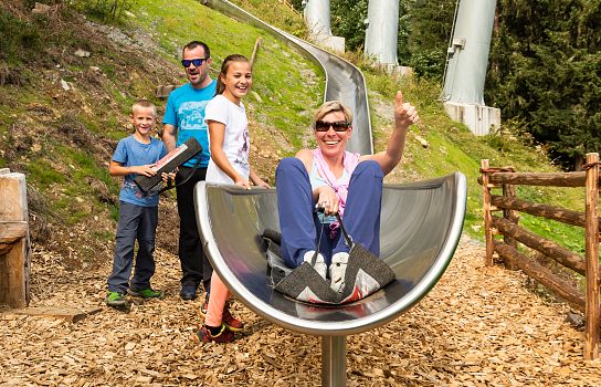 Vier Personen erfreuen sich an einer Sommerrodelbahn. Eine Frau sitzt lachend im Rodel, während zwei Kinder und ein Mann daneben stehen und zusehen.