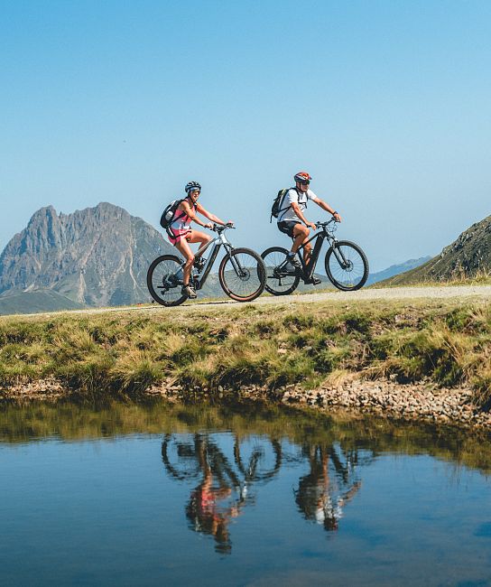 Zwei Radfahrer fahren auf einem schmalen Pfad in den Bergen neben einem klaren See. Im Hintergrund erhebt sich ein markanter Berggipfel unter blauem Himmel.