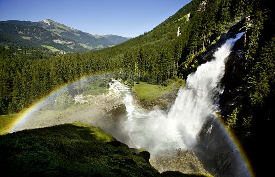 Eine beeindruckende Landschaft mit einem großen Wasserfall, umgeben von dichten Wäldern und Bergen. Ein Regenbogen spannt sich über das Tal.