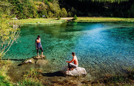 Zwei Menschen stehen auf Felsen an einem klaren, türkisblauen See in einer waldreichen Landschaft. Das Wasser spiegelt den grünen Wald und den blauen Himmel.