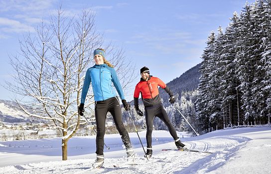 Zwei Personen beim Langlaufen auf einer Schneelandschaft, umgeben von schneebedeckten Bäumen und bergen. Sie tragen Winterkleidung in Blau und Rot.