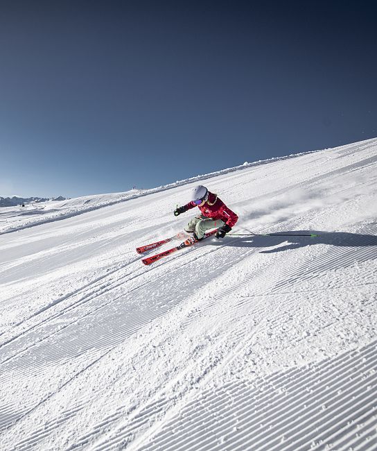 Ein Skifahrer in roter Kleidung fährt mit hoher Geschwindigkeit eine schneebedeckte Piste hinunter. Der Himmel ist klar und die Berge sind im Hintergrund sichtbar.