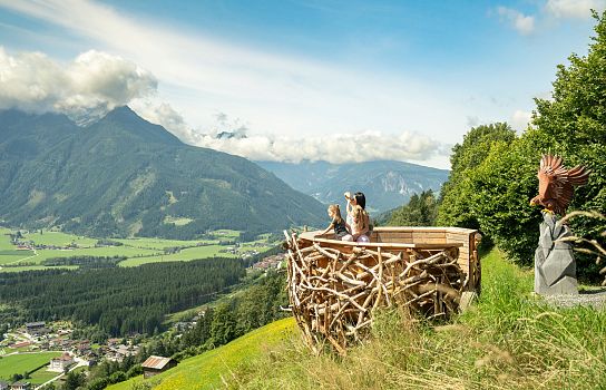 Holzplattform mit Ausblick auf ein alpines Tal, grüne Wälder und Dörfer. Im Hintergrund sind beeindruckende Berge unter einem blauen Himmel zu sehen.