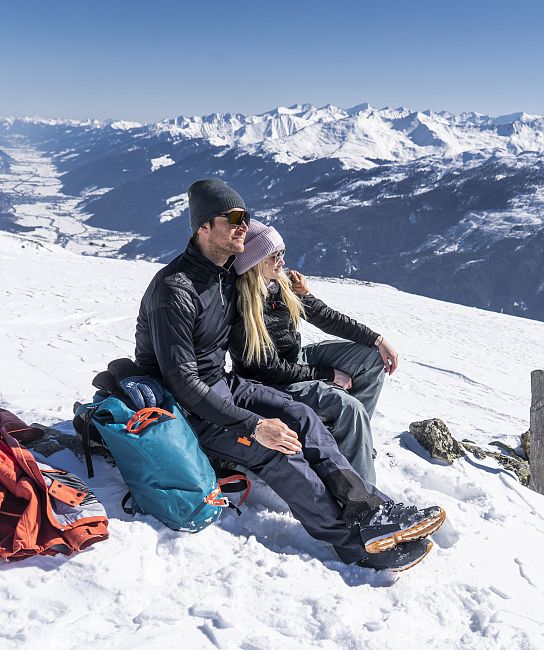 Ein Paar sitzt in Winterkleidung auf einem verschneiten Berghang mit Ausblick auf schneebedeckte Berge unter klarem, blauem Himmel.