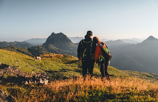 Ein Paar in Wanderkleidung steht auf einer grünen Bergwiese und blickt auf eine majestätische Berglandschaft im Sonnenuntergang.