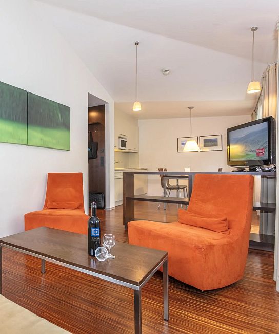 Modernes Wohnzimmer mit Holzboden, zwei orangefarbenen Sesseln, einem Couchtisch mit einer Weinflasche und Gläsern, sowie einem Flachbildfernseher an der Wand.