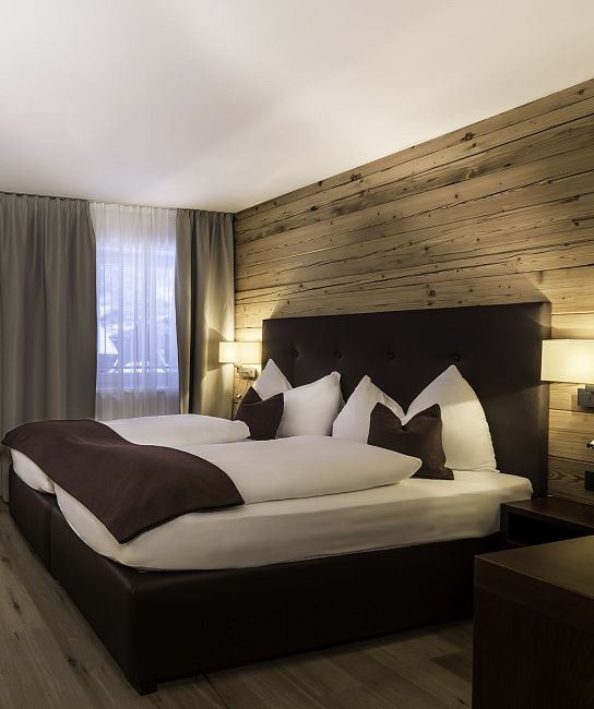 Ein modernes Hotelzimmer mit Doppelbett, Holzwand, zwei Sesseln und einem Flachbildfernseher. Große Fenster sorgen für viel Licht und eine einladende Atmosphäre.