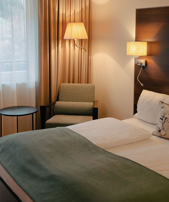 Ein Hotelzimmer mit einem Doppelbett, Holzverkleidung an den Wänden, großen Fenstern mit Vorhängen und bequemen Stühlen neben einem kleinen Tisch.