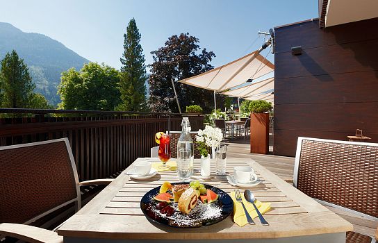 Ein gedeckter Tisch auf einer Sonnenterrasse mit einem Teller Essen, Getränken und Blick auf die Berge und Bäume im Hintergrund an einem sonnigen Tag.