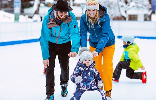 Familie beim Schlittschuhlaufen; Erwachsene unterstützen ein kleines Kind in bunter Winterkleidung auf dem Eis, während ein weiteres Kind im Hintergrund spielt.