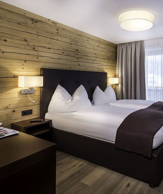 Gemütliches Hotelzimmer mit Doppelbett, Holzwand und moderner Einrichtung. Weiche Beleuchtung und Fenster mit Vorhängen sorgen für eine einladende Atmosphäre.