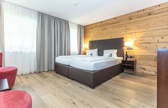 Gemütliches Hotelzimmer mit Holzwänden, zwei zusammengestellten Einzelbetten, großen Vorhängen, einem roten Sessel und warmer Beleuchtung.