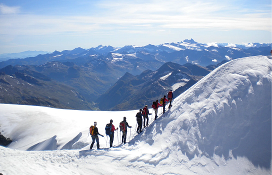 Eine Gruppe von Bergsteigern wandert in einer Schneelandschaft auf einem schmalen Pfad. Im Hintergrund sind verschneite Berge unter einem blauen Himmel sichtbar.